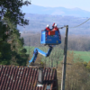 Two workers in a blue bucket lift repair power lines above a tiled roof in a rural valley setting.