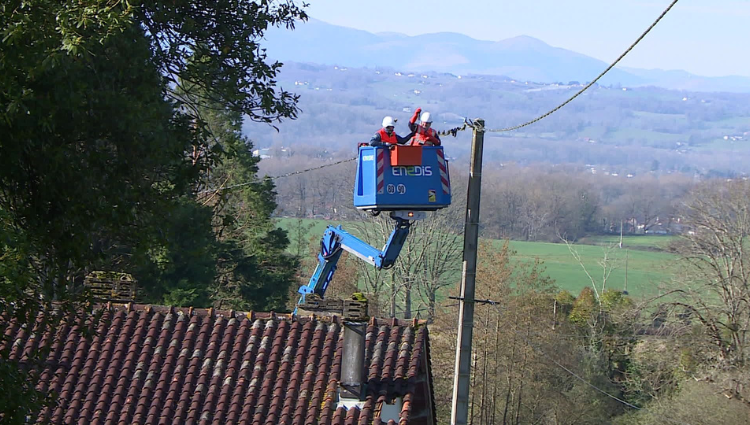 Two workers in a blue bucket lift repair power lines above a tiled roof in a rural valley setting.