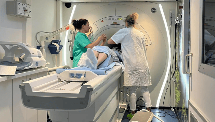 Patient on an examination table is assisted by two clinicians beside an MRI machine in a hospital room, preparing for scanning ground truth image context.