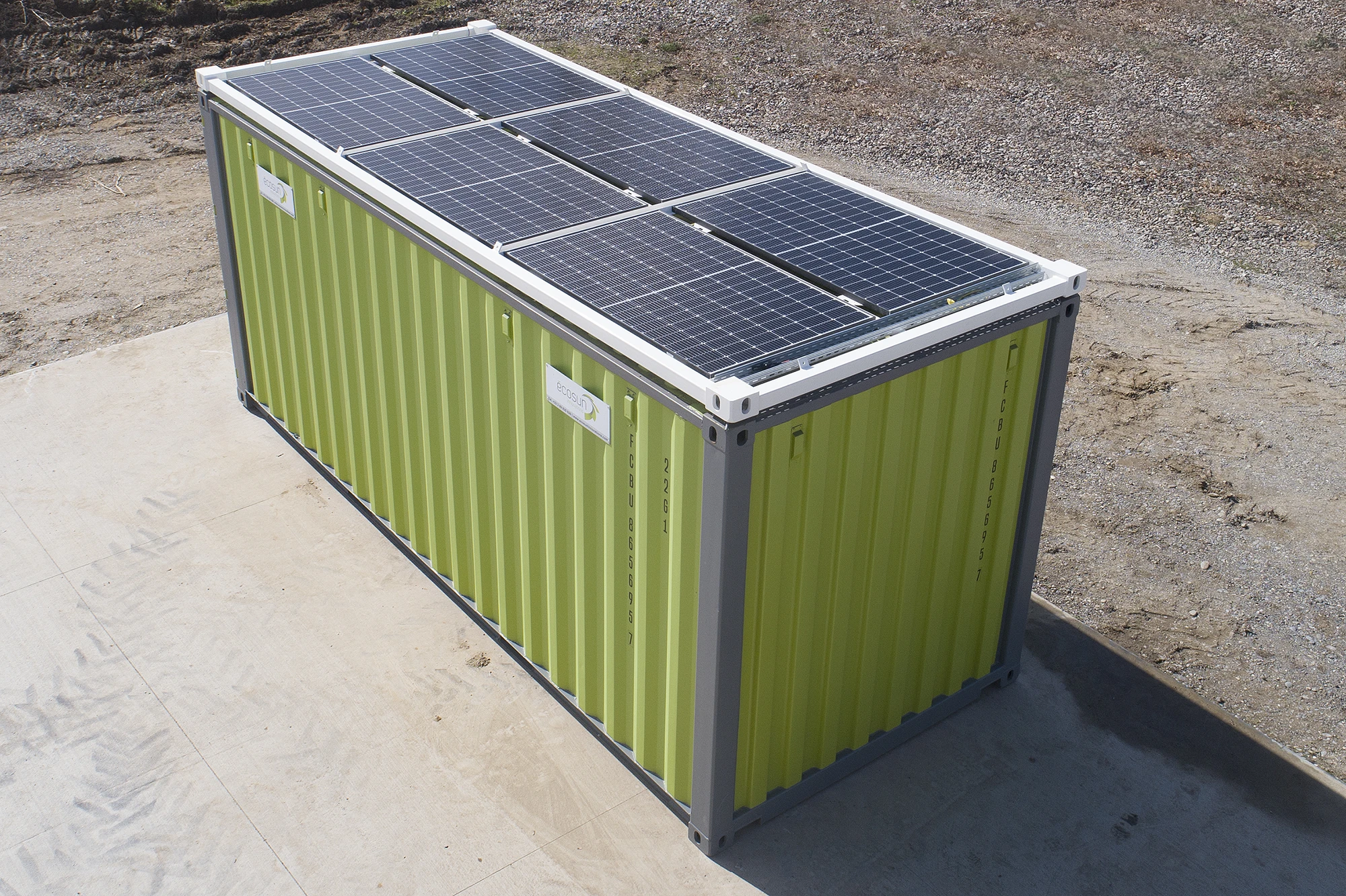 Green shipping container with solar panels mounted on top, placed outdoors on a construction site or barren yard.