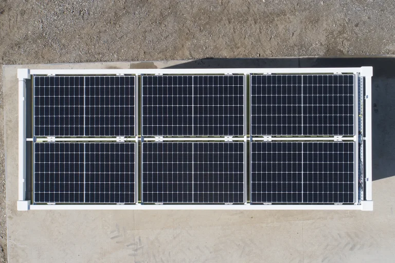 Top-down view of six solar panels arranged in two rows of three on a white frame atop a concrete pad in a dry, dusty area.